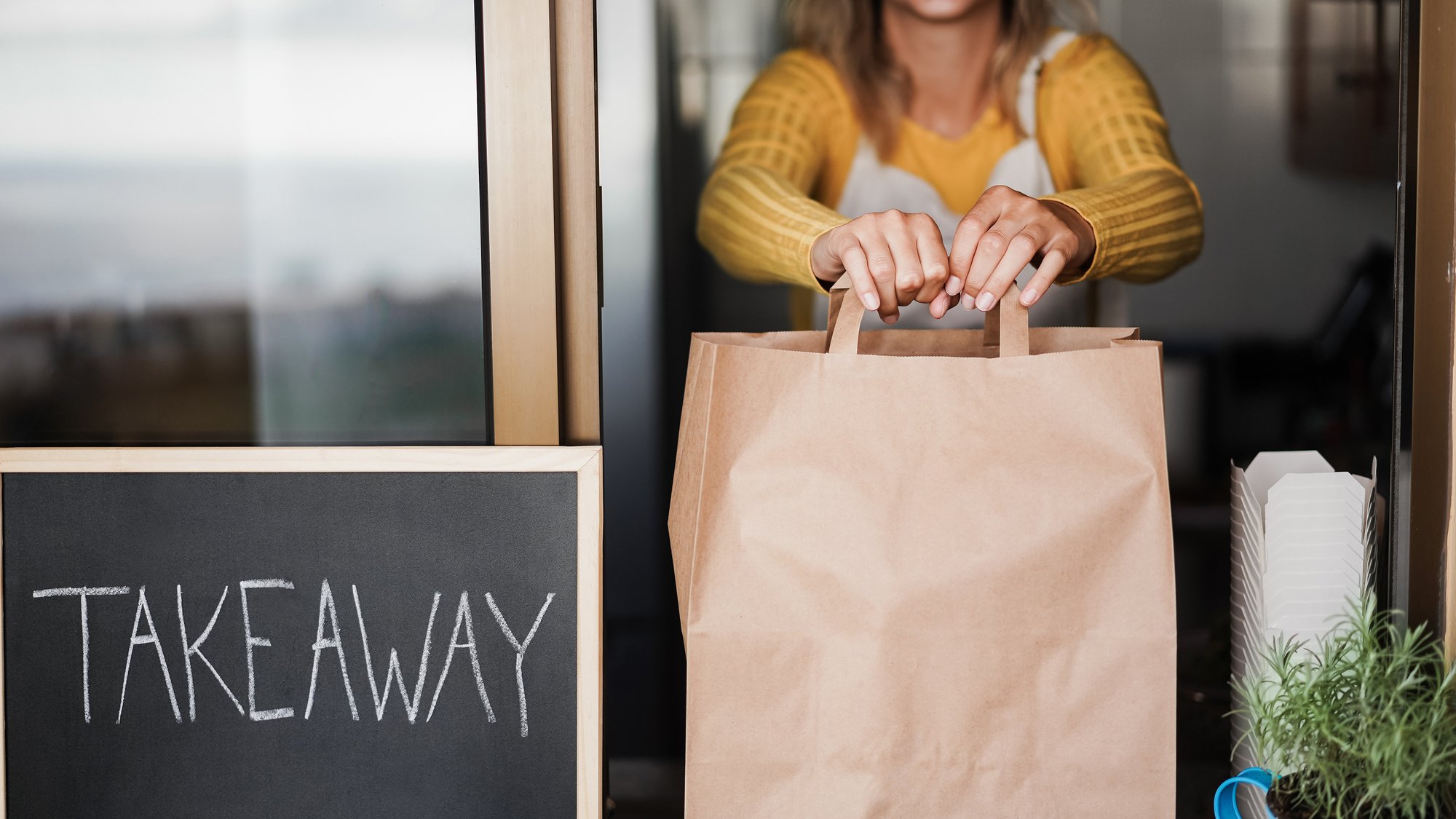 Young woman preparing takeaway food for fast delivery inside ghost kitchen - Focus on hands holding bag
